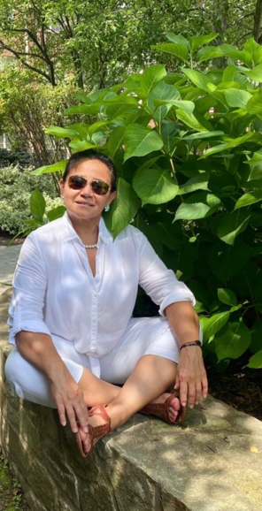 Woman meditating outdoors surrounded by lush green plants and trees.